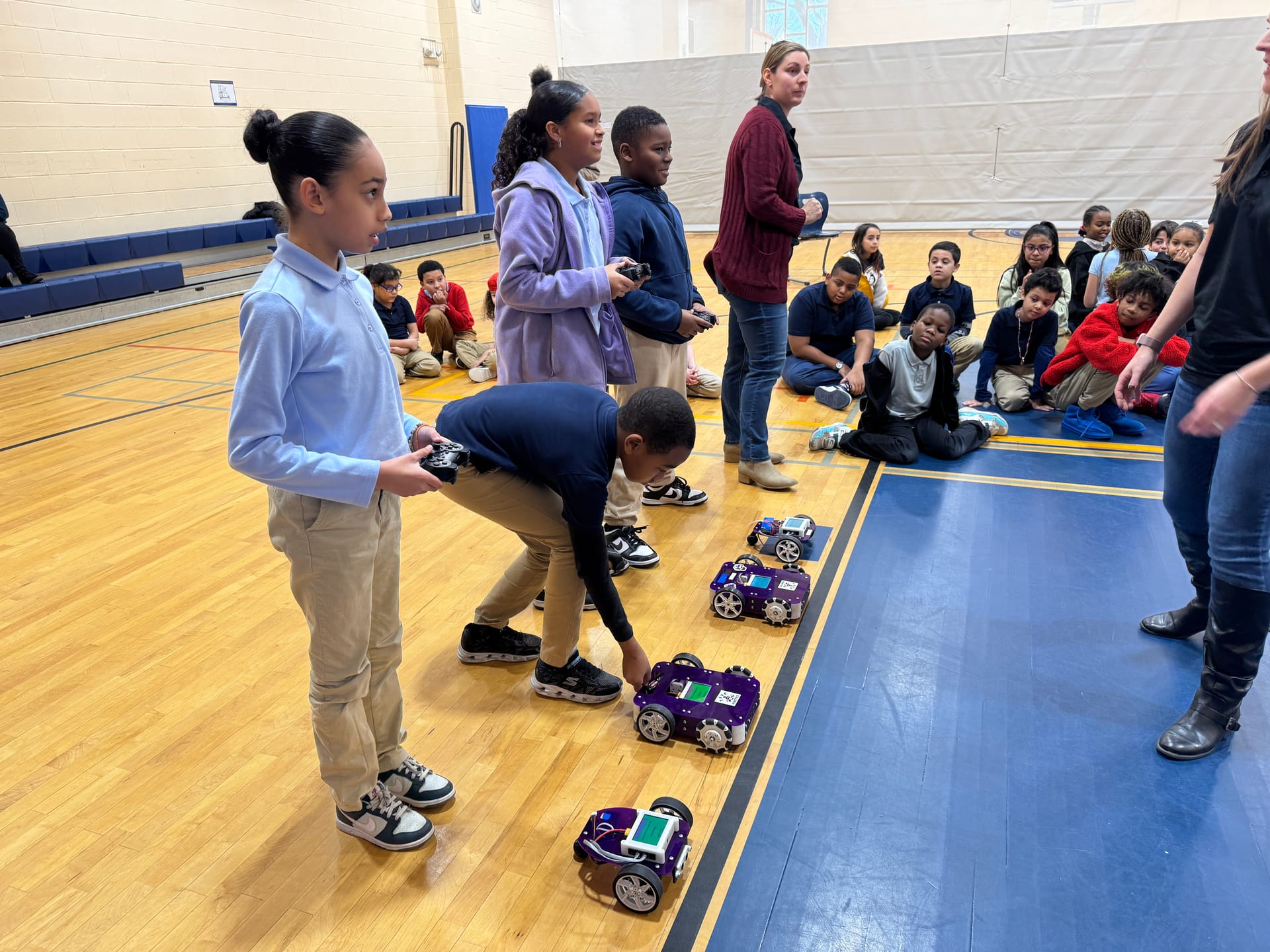 Four students hold game controllers while a classmate crouches to set a purple robot on the gym floor