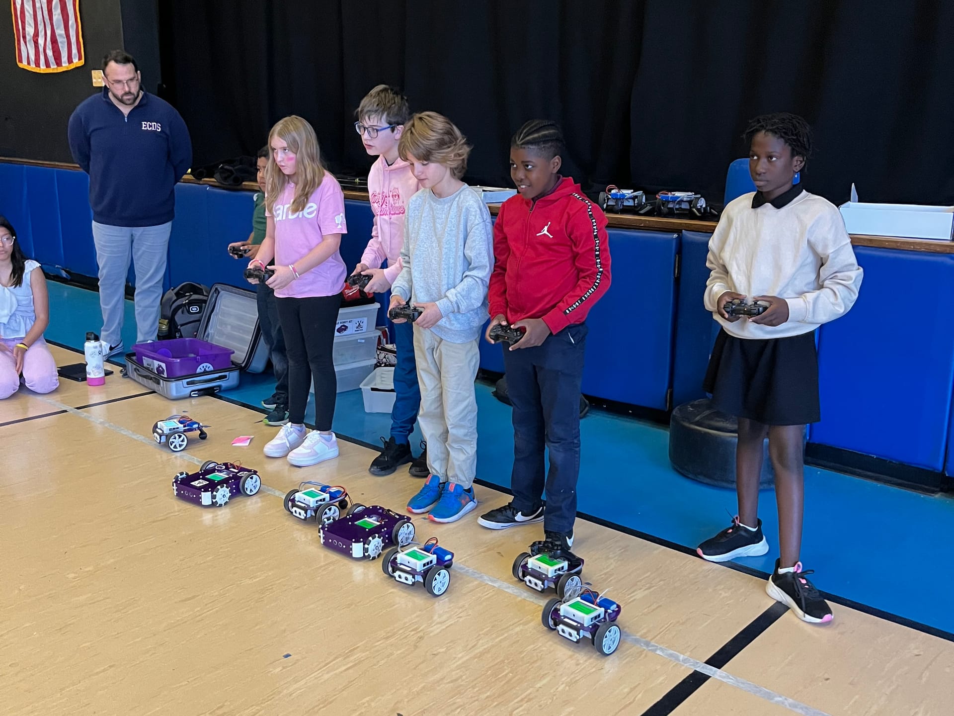 Five students stand in a row with game controllers driving Axle and Pilot robots across a gym floor as a teacher watches
