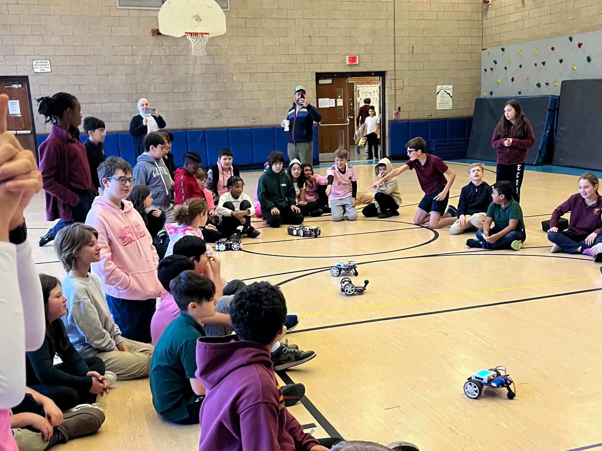 Students sit and stand around the edge of a gym floor and watch four robot kits drive between them