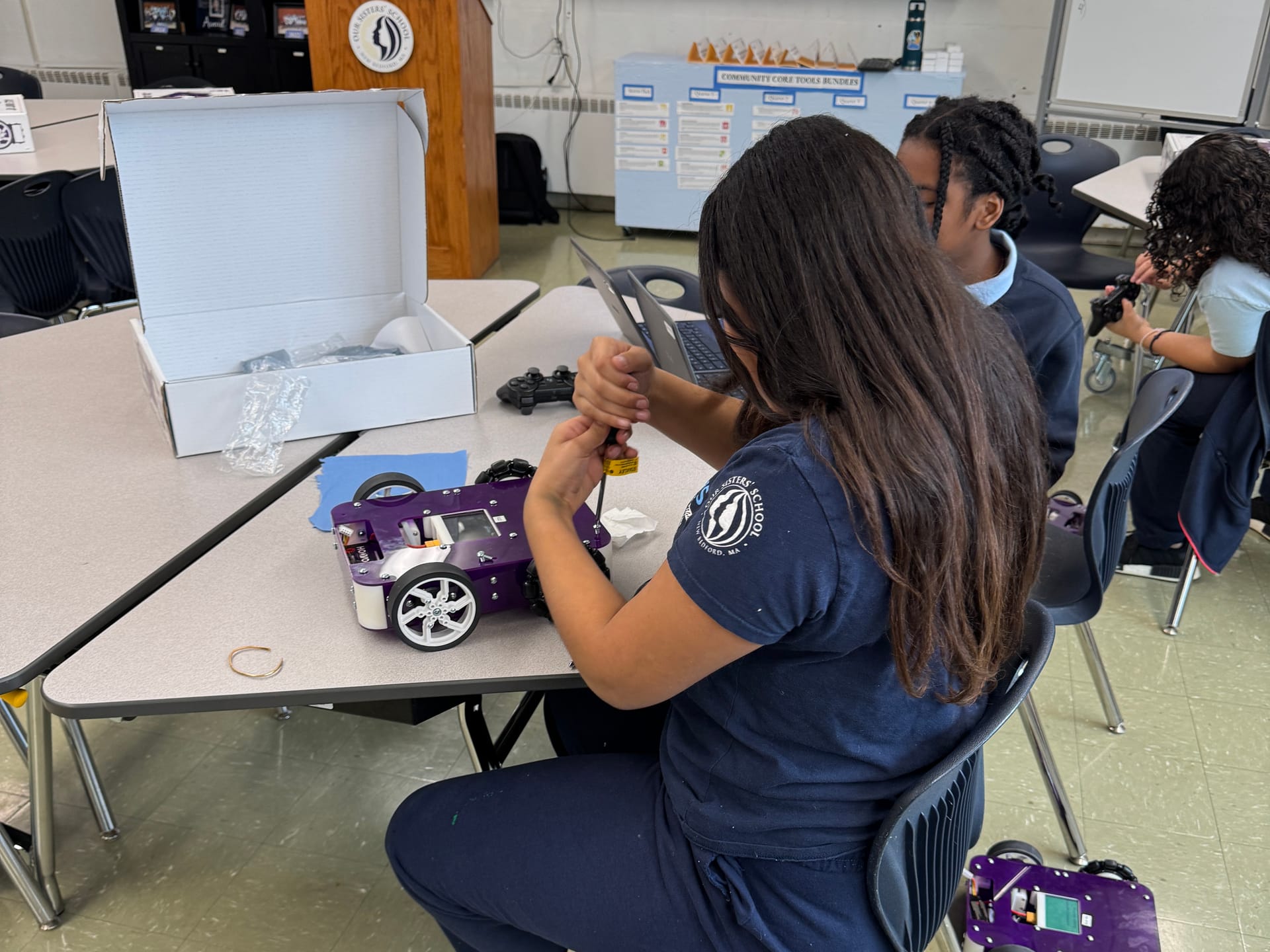 A middle school student uses a screwdriver to assemble an Axle robot at a classroom table with the kit box open beside it