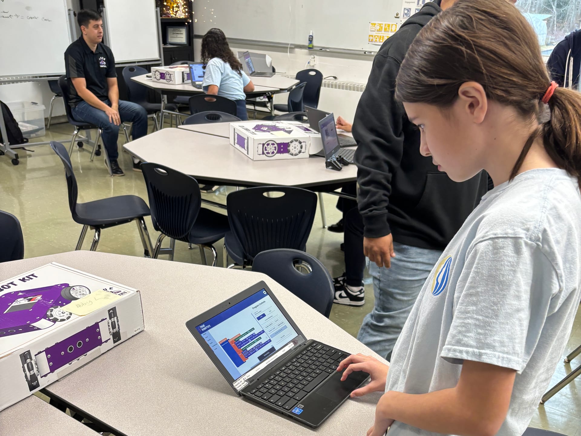 A middle school student writes a block program in True Robotics Playground on a Chromebook with an Axle Robot Kit box open beside her