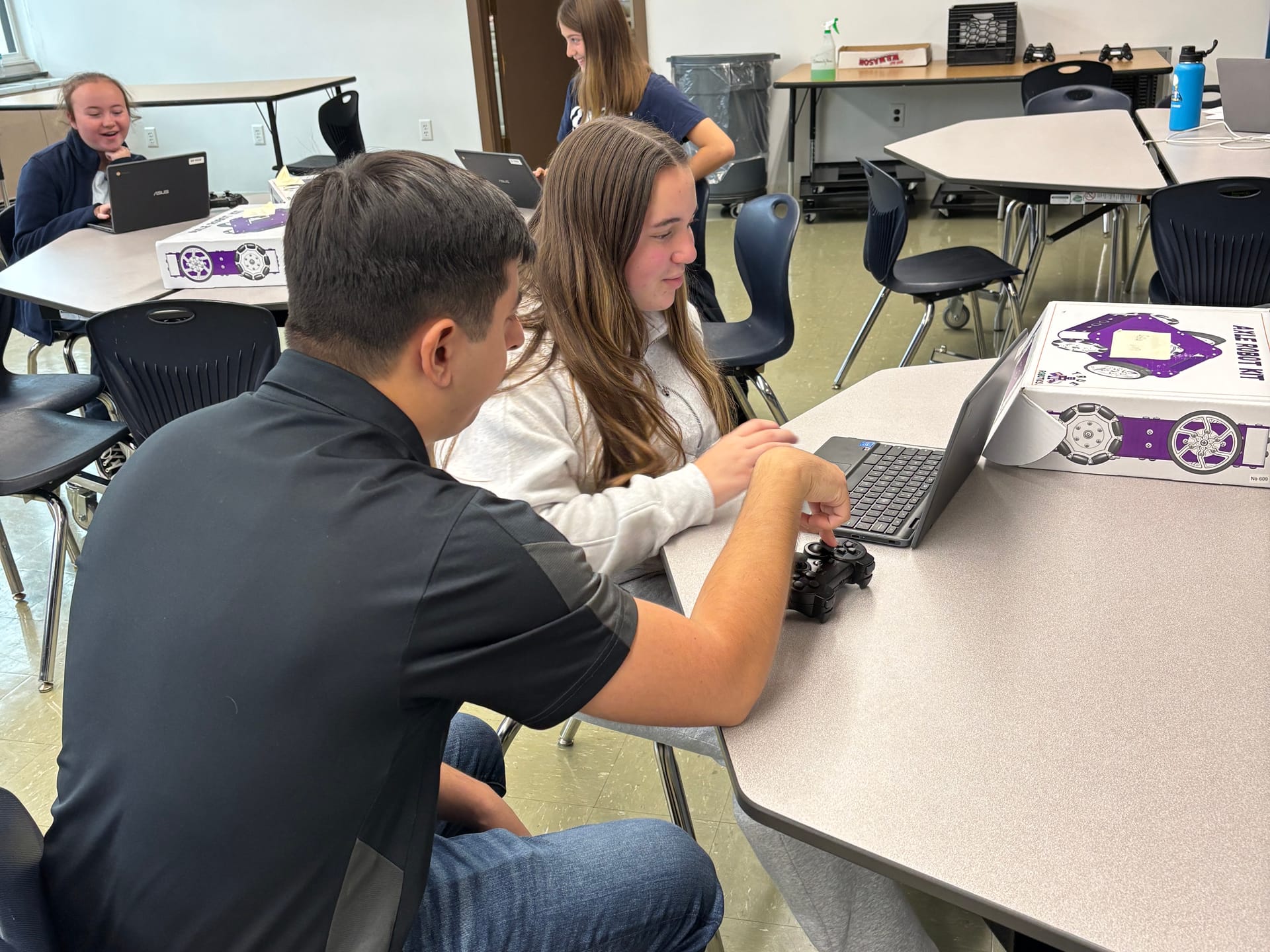 A True Robotics instructor sits with a middle school student at a Chromebook, game controller in hand, beside an Axle Robot Kit box