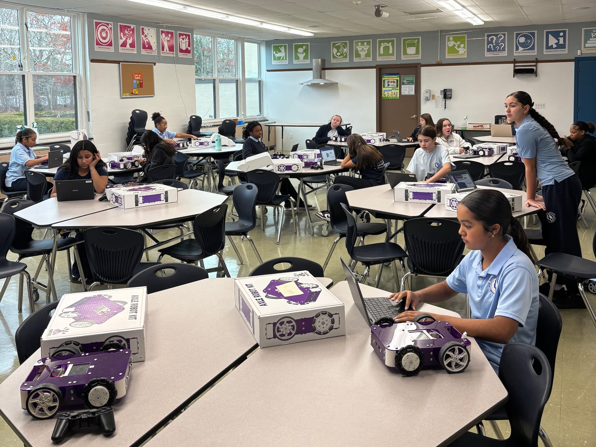 Students work at tables across a classroom with Axle Robot Kit boxes and Chromebooks while one student codes beside a purple Axle robot
