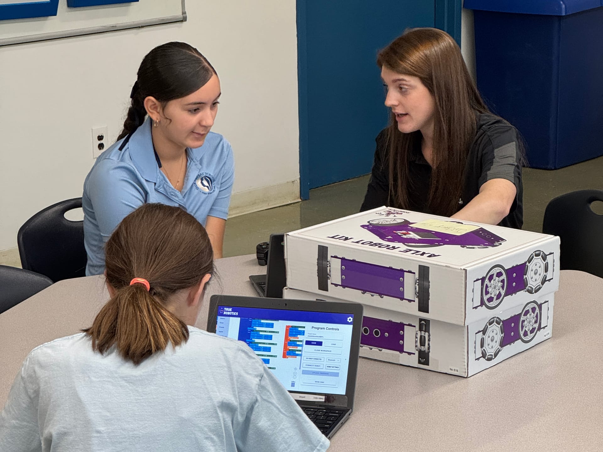 Three students work around a table with an Axle Robot Kit box beside them while one of them builds a program on a laptop