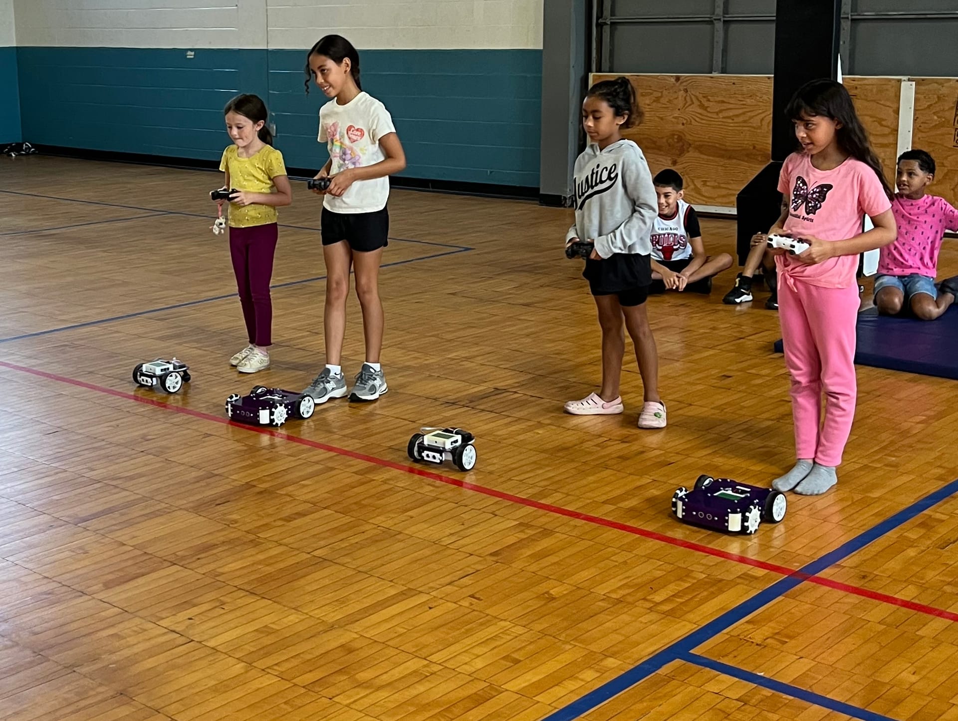Four students with game controllers drive purple robot kits in a line on a gym floor