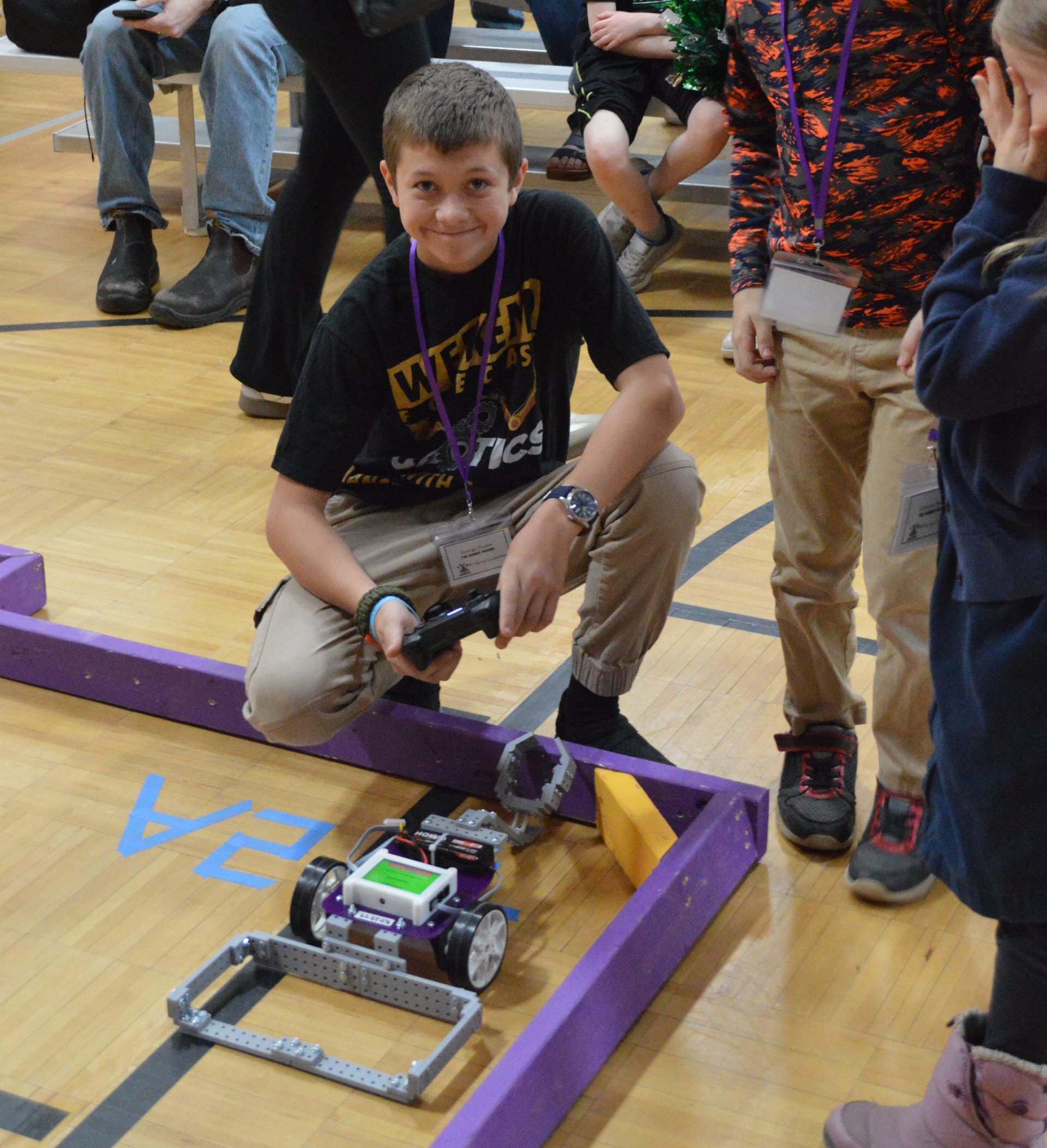 A student kneels at the edge of a purple course with a game controller and grins while driving a robot built with gray FlexForge beams