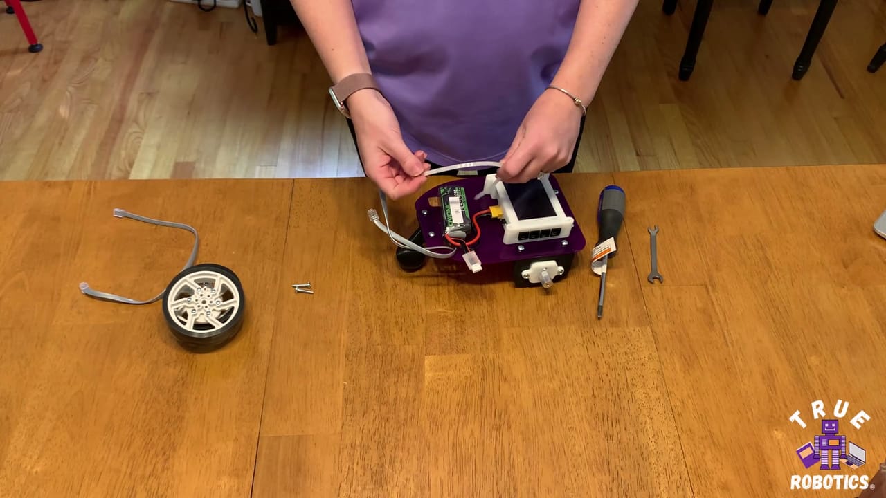 Hands fit a motor to a part built Pilot Robot Kit on a wood table beside a wheel and a screwdriver